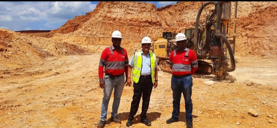Dr Hegde in a hard hat and safety vest with two colleagues at a limestone quarry, a drilling rig behind them